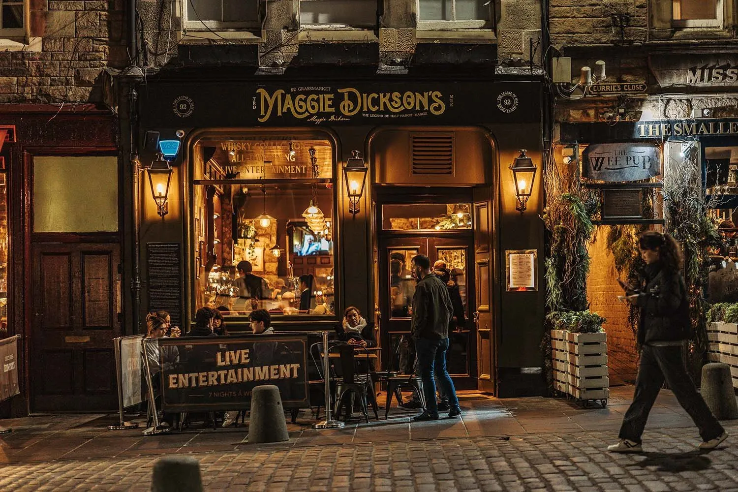 This street-level shot captures the inviting facade of "Maggie Dickson's," a traditional pub glowing warmly at night amidst historic stone buildings and a cobblestone street. The dark green exterior, adorned with gold lettering and ornate wall lanterns, frames large windows revealing a bustling interior with patrons, a well-lit bar, and ambient pendant lighting. Outdoor seating with a "LIVE ENTERTAINMENT" sign adds to the lively, welcoming atmosphere, enhanced by the soft, amber glow emanating from within.