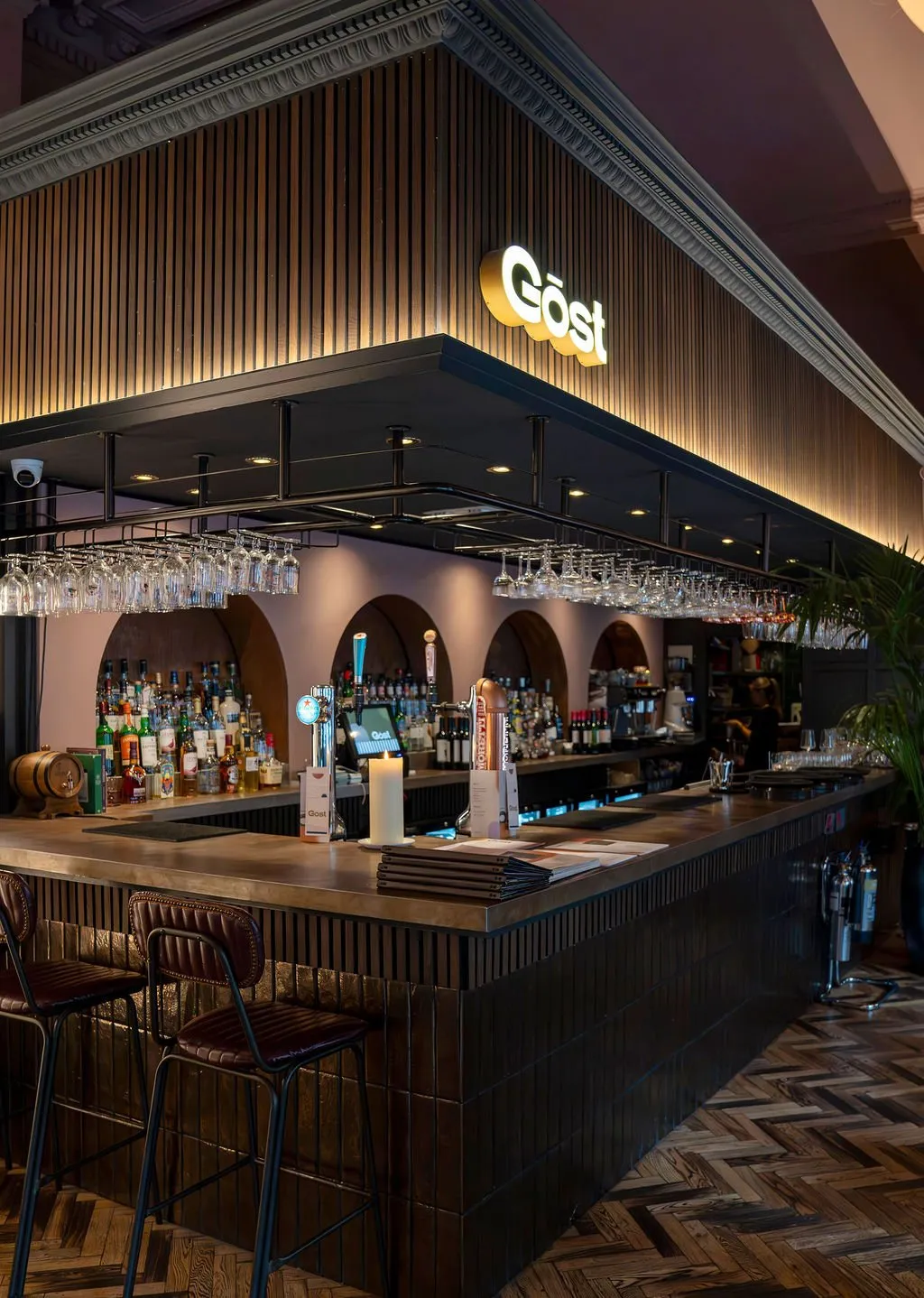 This sophisticated bar interior features a long, dark-tiled and slatted counter topped with a rich, dark surface, complemented by elegant brown leather bar stools. The back bar showcases arched niches in a soft pink plaster, brightly illuminating a vast selection of bottles, while an overhead black metal gantry holds an array of glassware. Warm uplighting from behind vertical wooden slats on the bar canopy and recessed spotlights create an inviting, intimate atmosphere, contrasting with the ornate original ceiling molding and a modern herringbone wood floor.