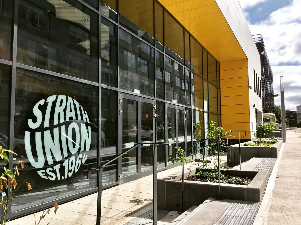 This image captures the vibrant, modern exterior of a university union building, featuring a striking facade dominated by expansive dark-framed glass panels and a bold, bright yellow upper cladding. The entrance is flanked by modular concrete planters with sparse greenery and integrated steps, creating a functional yet inviting approach. The bright daylight illuminates the scene, highlighting the reflective quality of the glass and the clean lines of the contemporary architecture.