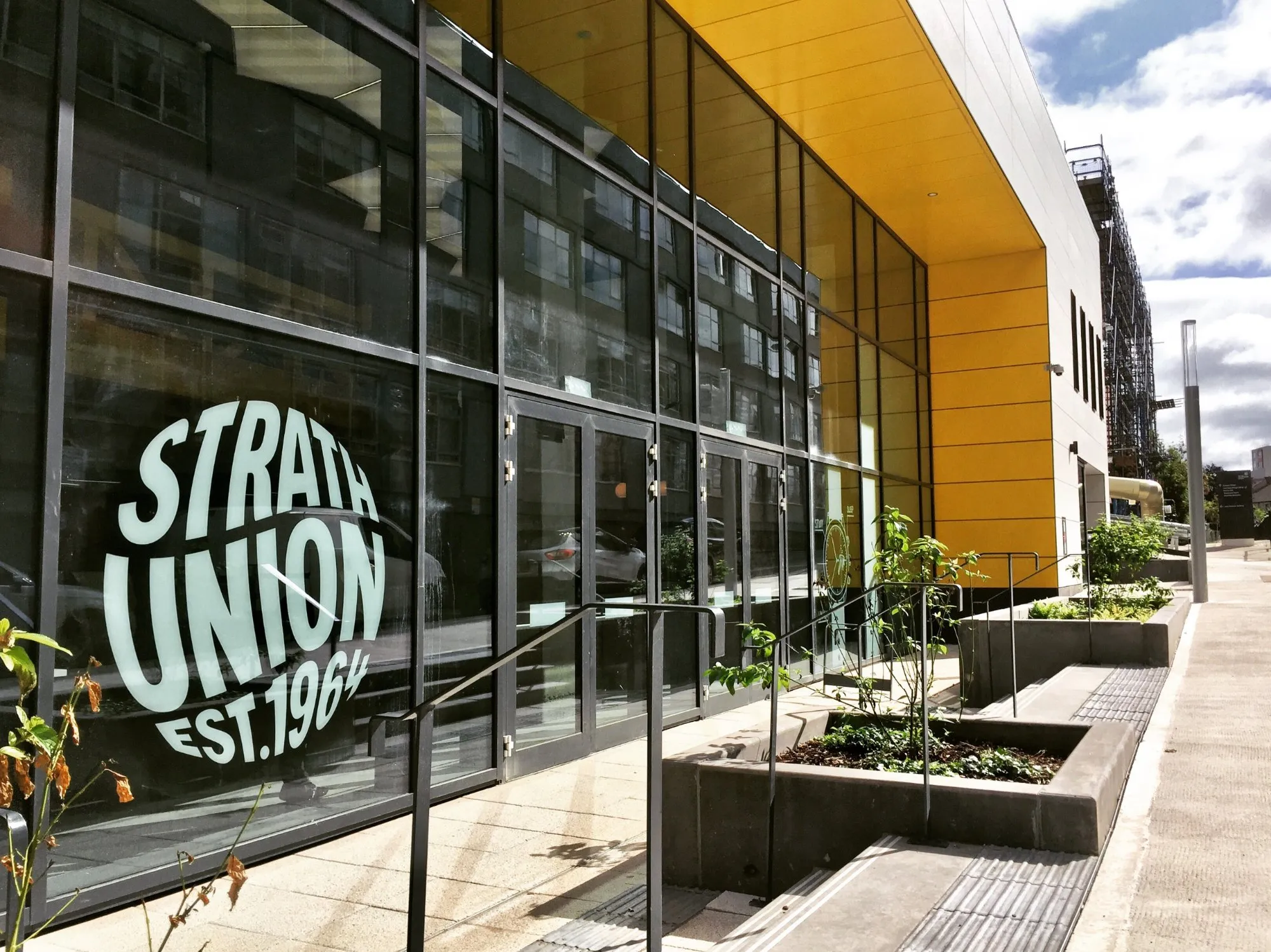 This image captures the vibrant, modern exterior of a university union building, featuring a striking facade dominated by expansive dark-framed glass panels and a bold, bright yellow upper cladding. The entrance is flanked by modular concrete planters with sparse greenery and integrated steps, creating a functional yet inviting approach. The bright daylight illuminates the scene, highlighting the reflective quality of the glass and the clean lines of the contemporary architecture.
