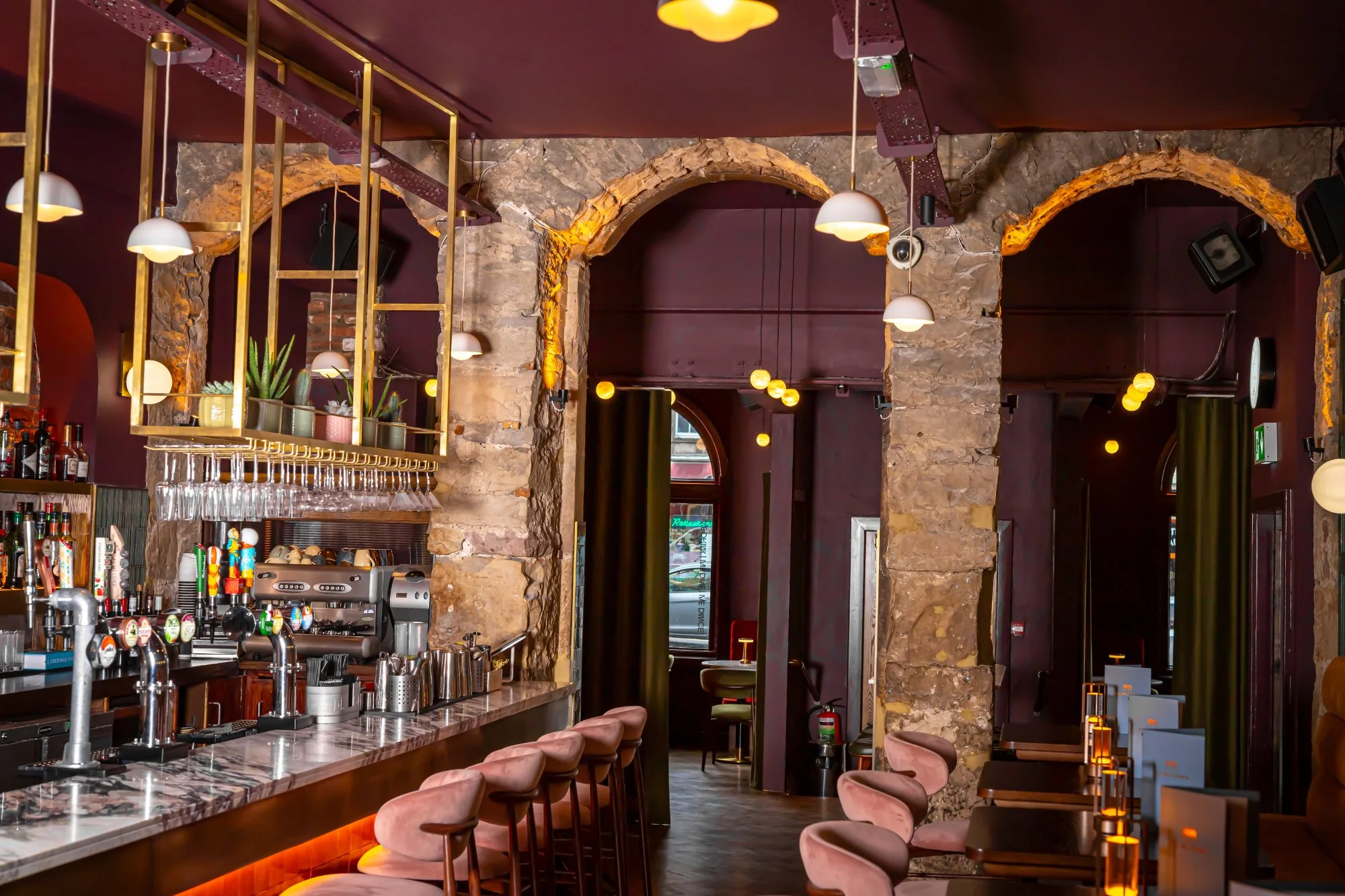 This vibrant hospitality space features a long, inviting bar counter with a polished marble top, complemented by plush pink velvet bar stools. Overhead, a striking brass gantry integrates both decorative plants and practical glass storage, illuminated by warm pendant lights. Beyond the bar, exposed rough-hewn stone arches frame a dining area with dark wood tables and a mix of pink and deep green velvet chairs, all set against rich plum-coloured walls and ceiling, creating an intimate and sophisticated atmosphere.