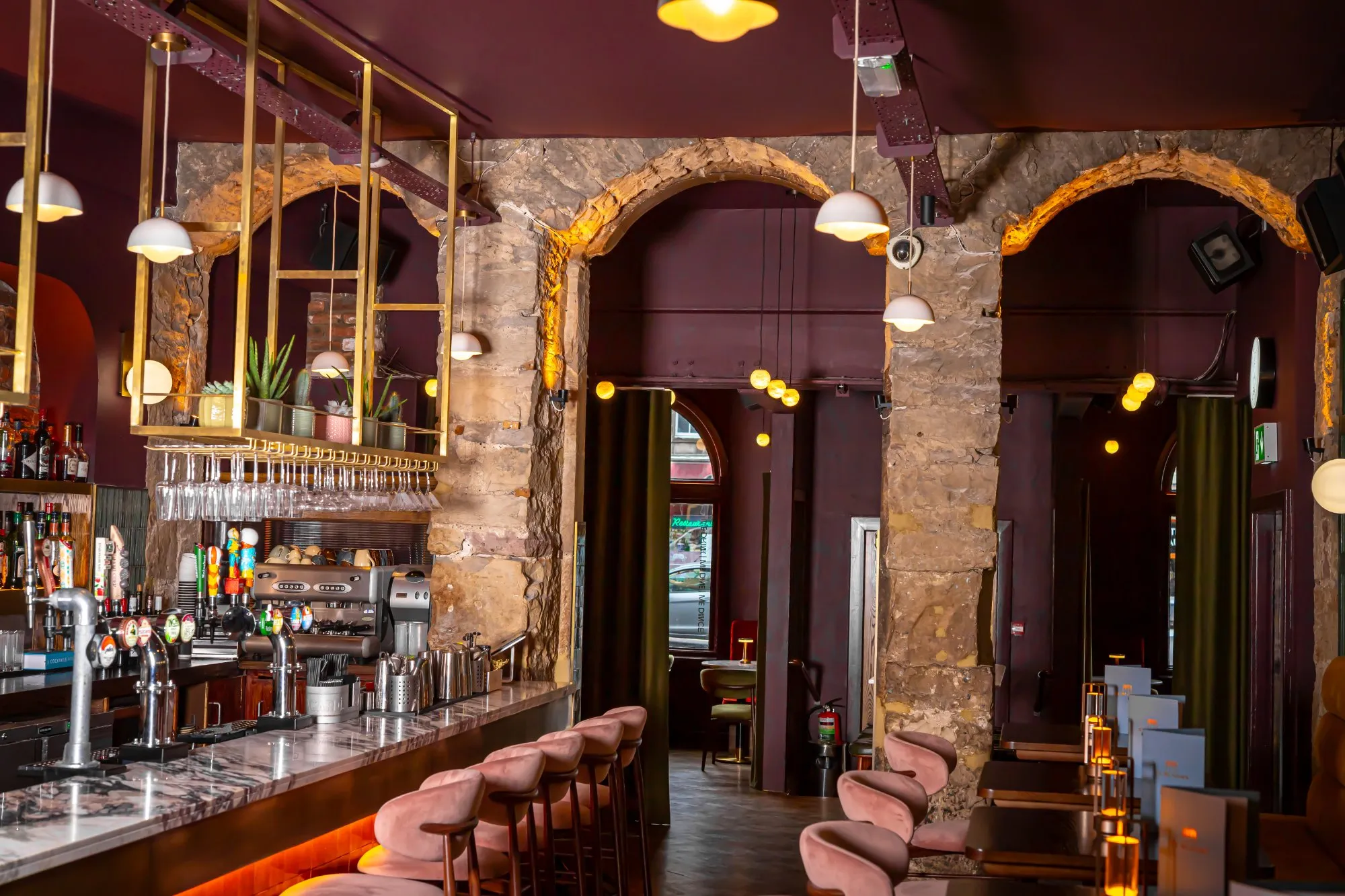 This vibrant hospitality space features a long, inviting bar counter with a polished marble top, complemented by plush pink velvet bar stools. Overhead, a striking brass gantry integrates both decorative plants and practical glass storage, illuminated by warm pendant lights. Beyond the bar, exposed rough-hewn stone arches frame a dining area with dark wood tables and a mix of pink and deep green velvet chairs, all set against rich plum-coloured walls and ceiling, creating an intimate and sophisticated atmosphere.