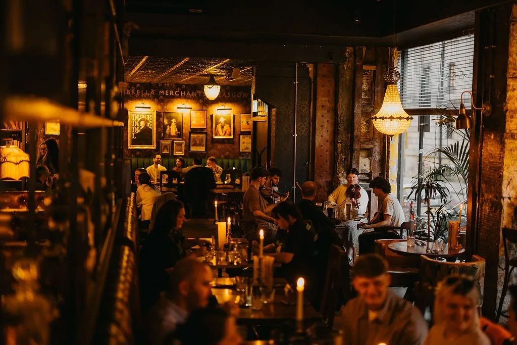 This atmospheric hospitality space features a rich, eclectic design, bathed in warm, low light from candles, wall sconces, and an ornate chandelier. Dark wood tables and deep green upholstered banquettes create intimate seating zones, complemented by an extensive gallery wall of framed art and a textured pressed-tin ceiling. The mood is lively and cozy, enhanced by live musicians and a bustling crowd, suggesting a vibrant evening venue with a vintage, speakeasy-inspired charm.