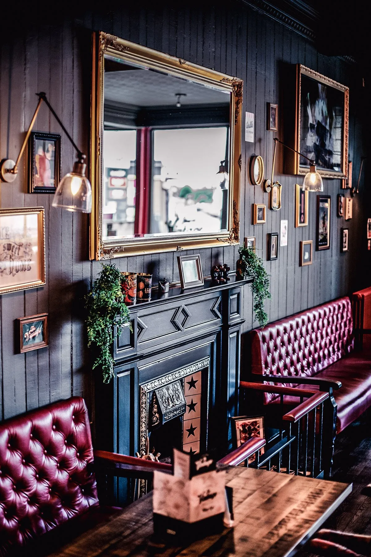 This intimate interior features dark wood paneling on the walls, creating a rich, moody backdrop for a gallery of framed art and an ornate gold-framed mirror above a classic dark fireplace mantel. Plush, deep red tufted leather banquettes and chairs offer comfortable seating around dark wooden tables, illuminated by warm industrial-style wall sconces. The space exudes a cozy, traditional pub or restaurant atmosphere, blending classic elements with a touch of eclectic charm.