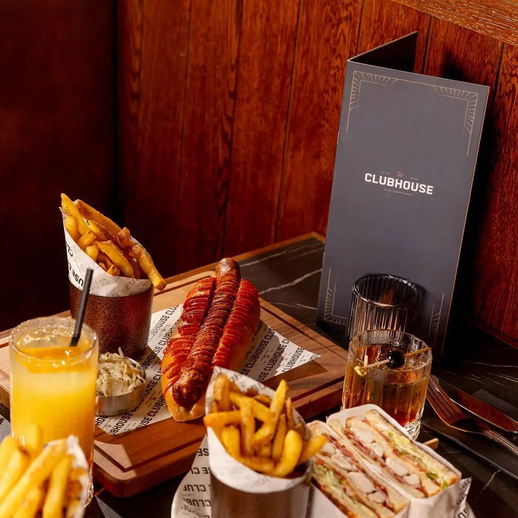 This close-up captures a sophisticated yet casual dining experience within a warmly lit interior. A dark veined marble tabletop grounds an array of classic comfort food – a gourmet hot dog, crispy fries, and a club sandwich – served on a wooden board and in stylish metal cups. The backdrop features rich, dark wood paneling, creating a cozy, club-like atmosphere, further enhanced by the warm, directional lighting that highlights the food and drinks, including an Old Fashioned and a vibrant orange juice. A chic menu with Art Deco accents completes the inviting scene.