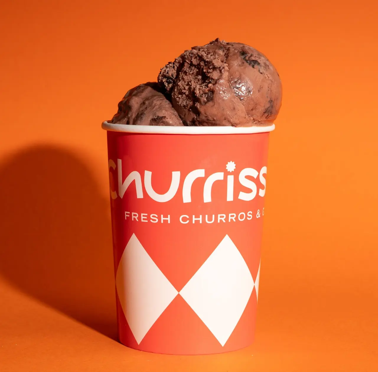 This is a striking product shot featuring two generous scoops of dark chocolate ice cream, visibly rich with chocolate chunks, served in a branded 'Churriss' paper cup. The cup itself is a vibrant red with a crisp white logo and a playful harlequin pattern on its lower half, stating 'Fresh Churros &'. The entire presentation is set against a bold, solid orange background, creating a high-contrast, energetic, and modern aesthetic, perfect for a dessert or fast-casual brand's promotional material.