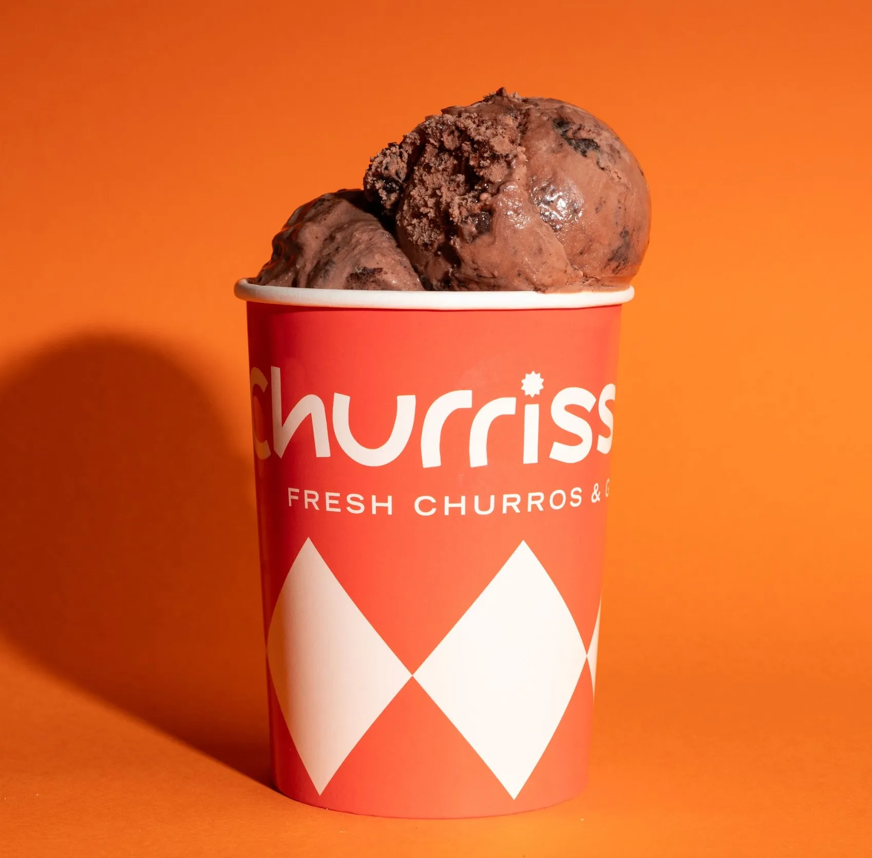 This is a striking product shot featuring two generous scoops of dark chocolate ice cream, visibly rich with chocolate chunks, served in a branded 'Churriss' paper cup. The cup itself is a vibrant red with a crisp white logo and a playful harlequin pattern on its lower half, stating 'Fresh Churros &'. The entire presentation is set against a bold, solid orange background, creating a high-contrast, energetic, and modern aesthetic, perfect for a dessert or fast-casual brand's promotional material.