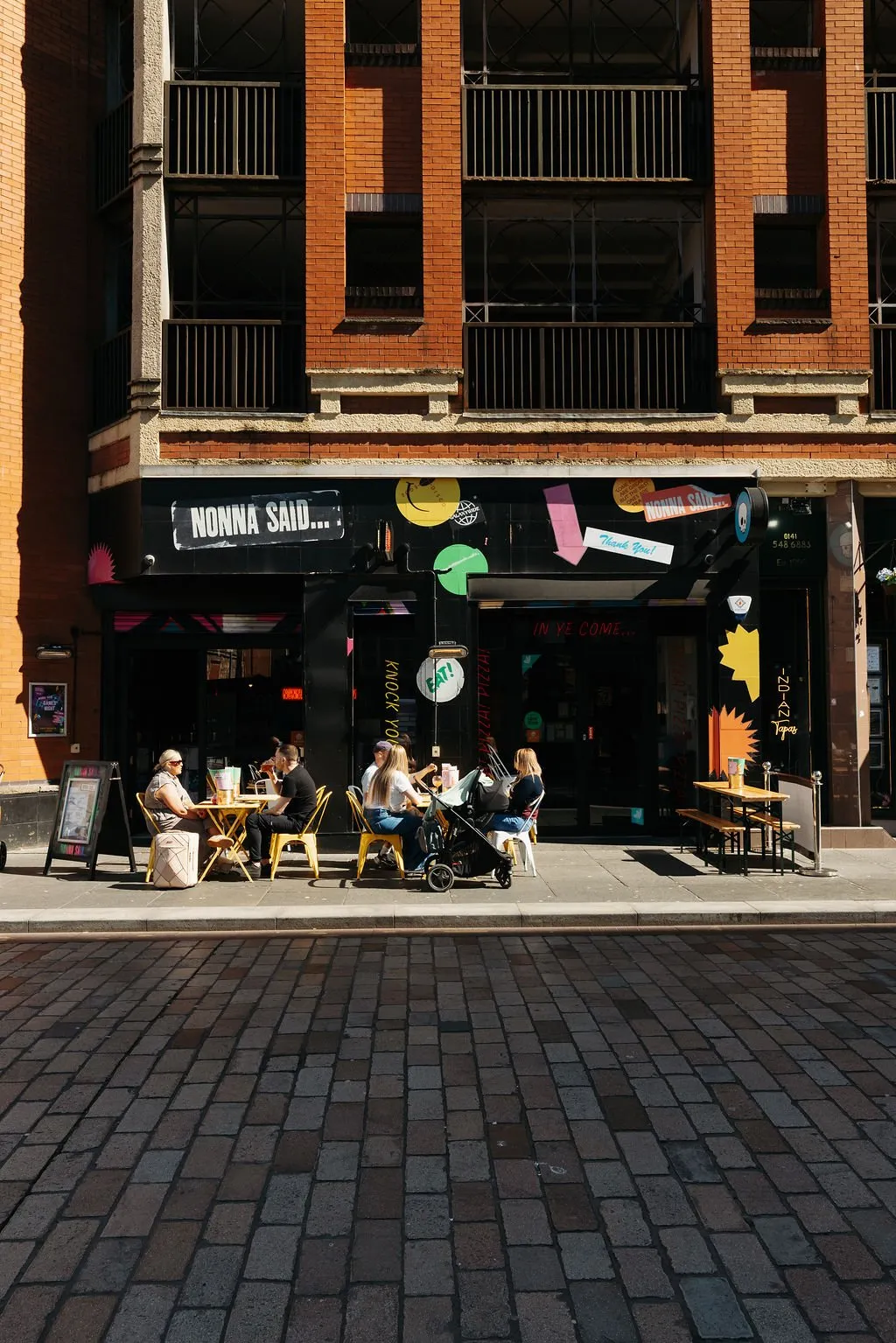 This is an exterior shot of a vibrant, urban cafe or restaurant, featuring a bold black storefront adorned with playful, colorful graphic stickers and signage. Outdoor seating on the pavement includes bright yellow metal chairs and tables, alongside more rustic wooden picnic benches, creating a casual and inviting atmosphere under bright daylight. The building above is a traditional red brick structure with dark metal balcony railings, contrasting with the modern, graphic-heavy ground floor.