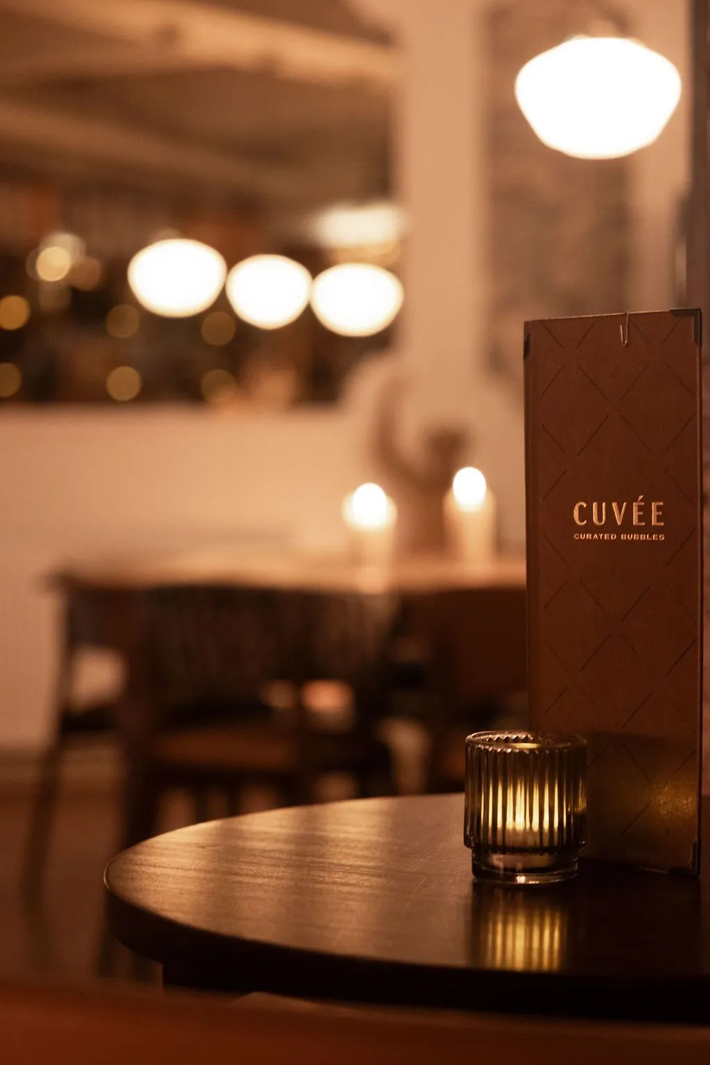 This intimate shot captures a sophisticated evening ambiance in a restaurant or bar, focusing on a dark, round wooden table. A ribbed glass votive holder casts a warm glow, illuminating a branded "CUVÉE" menu with a subtle diamond pattern. The blurred background reveals additional tables and soft, diffused overhead lighting, creating a cozy and inviting atmosphere perfect for a relaxed dining or lounge experience.
